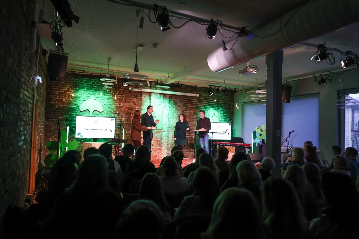 Paneldebatt på scenen med grønt lys og teglvegger, sett bakfra over publikum
