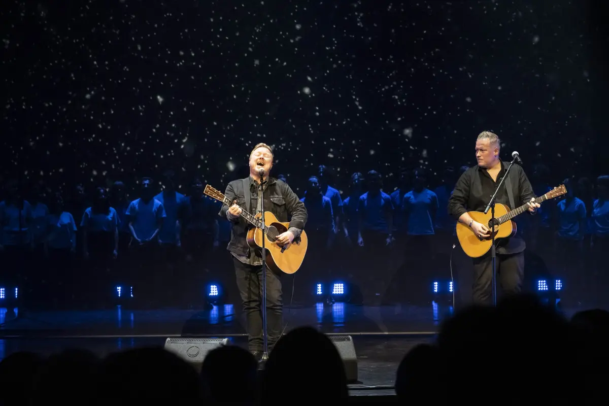 To gitarister på scenen foran stjerneteppe med ungdomskoret i blått lys bak
