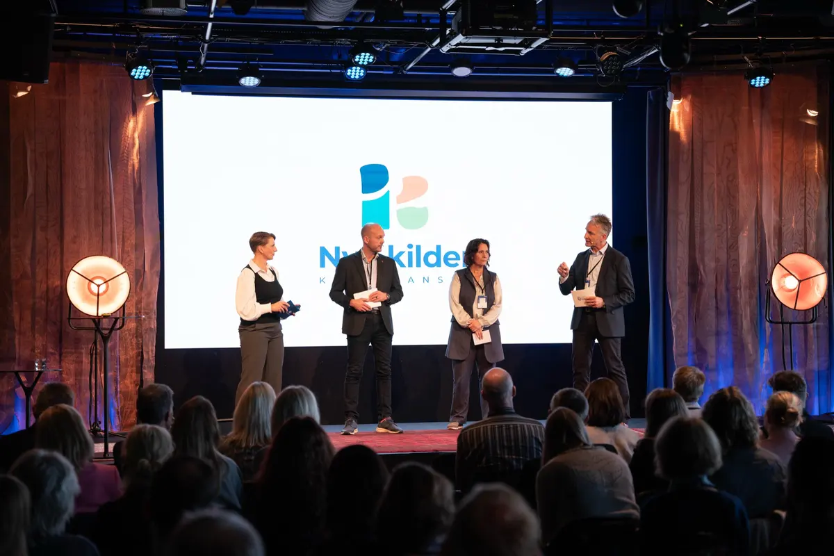 Fire personer står på scenen foran Nye kilder-skjermen mens publikum ser på