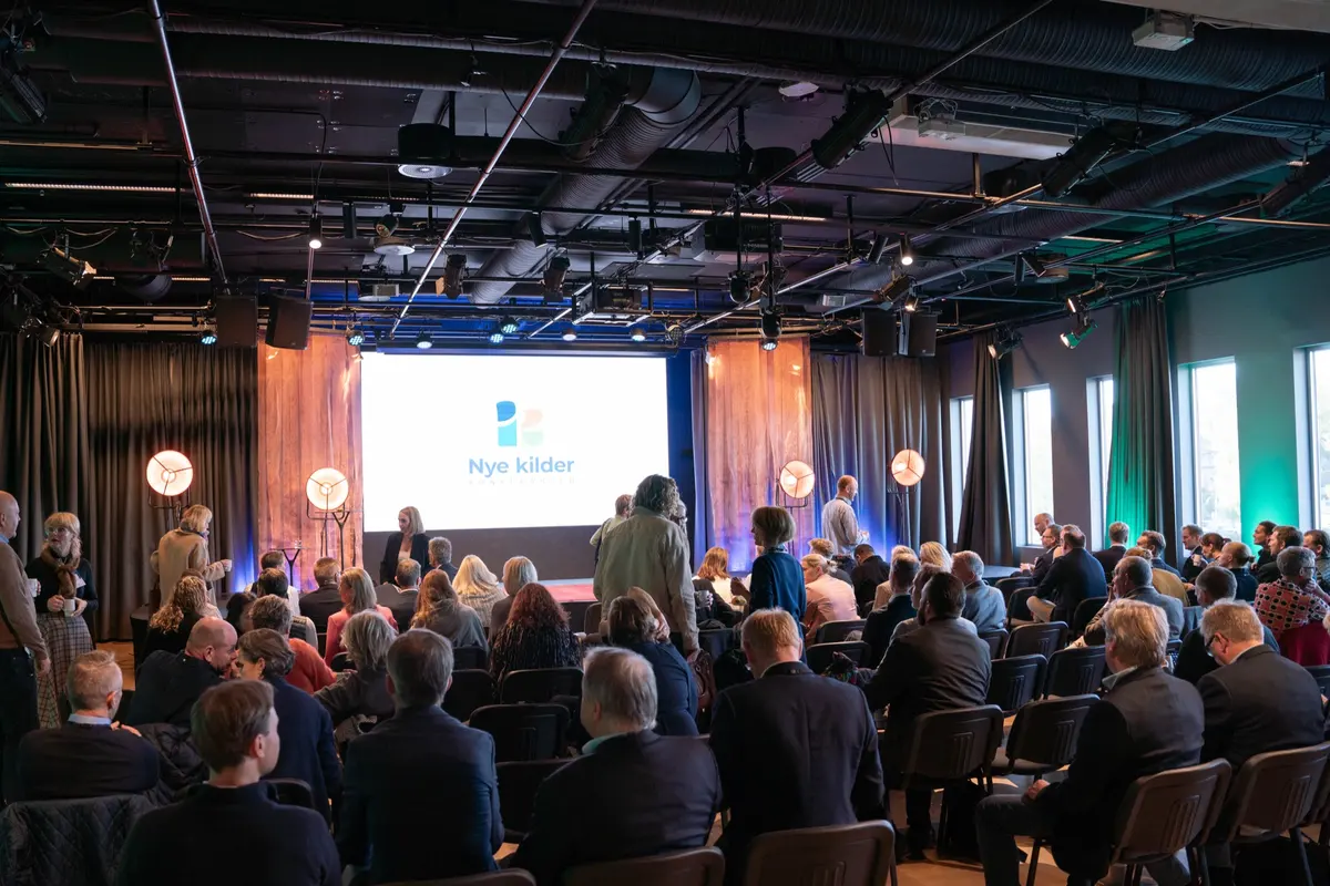 Oversikt over konferansesalen med publikum på stoler og Nye kilder-logo på skjermen