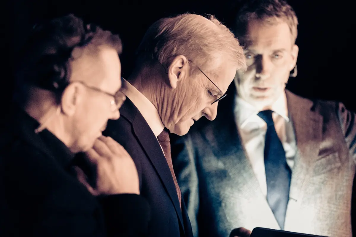Tre menn i dress ser ned på en skjerm i mørket backstage