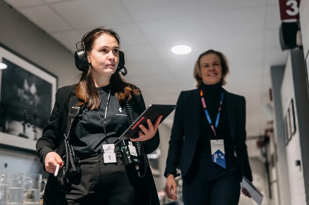 Backstage: to medarbeidere med akkreditering i korridoren, den ene med headset og nettbrett