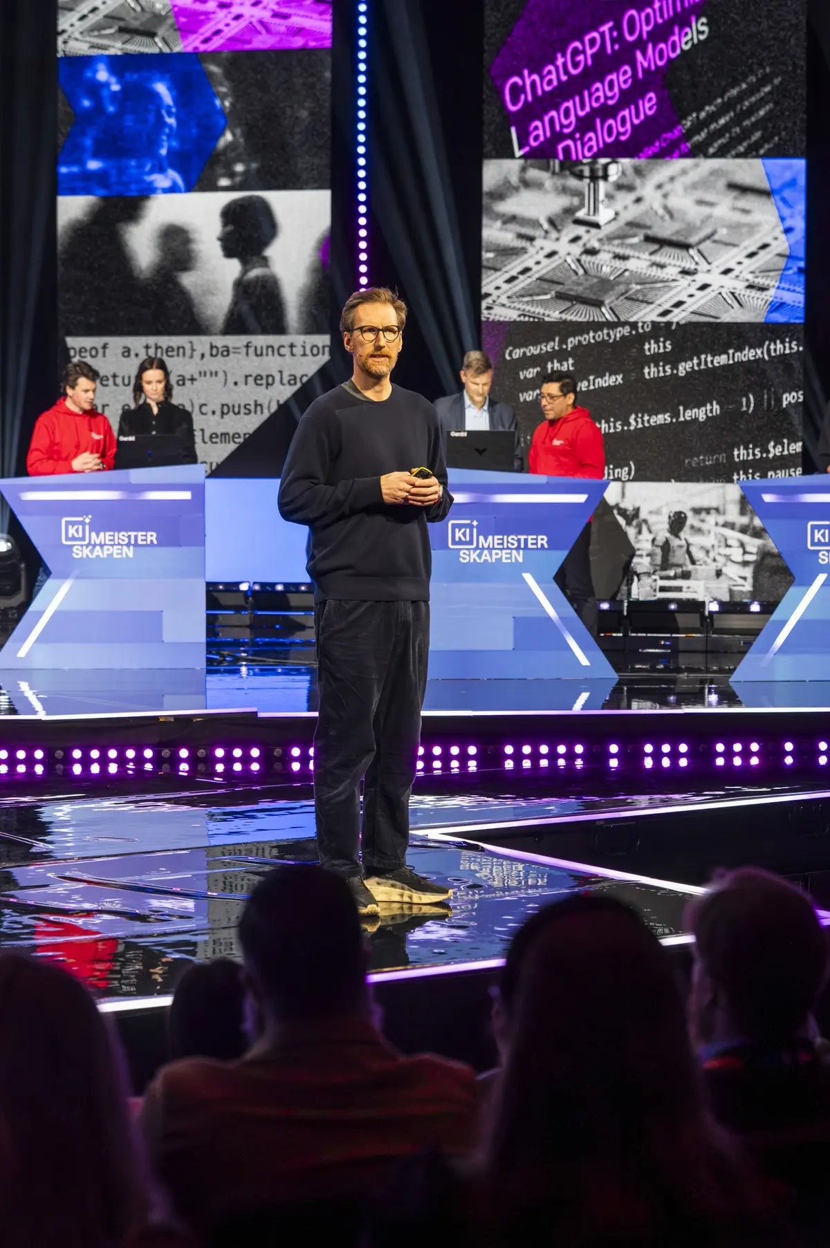 Taler midt på scenen med to lag ved pulter bak seg